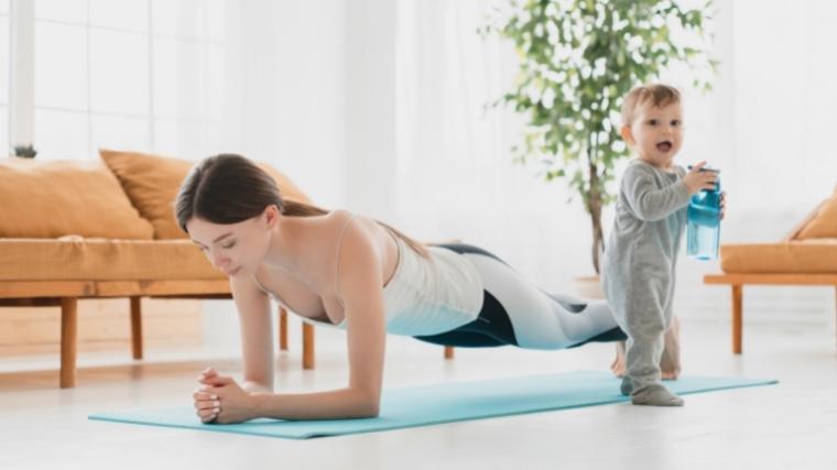 Woman performing a plank with a small child standing next to her holding a bottle