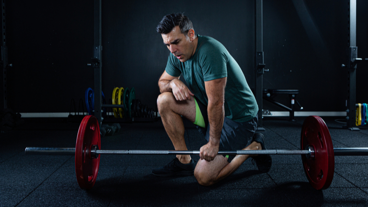 older man in dark gym kneeling on ground while holding barbell and breathing hard